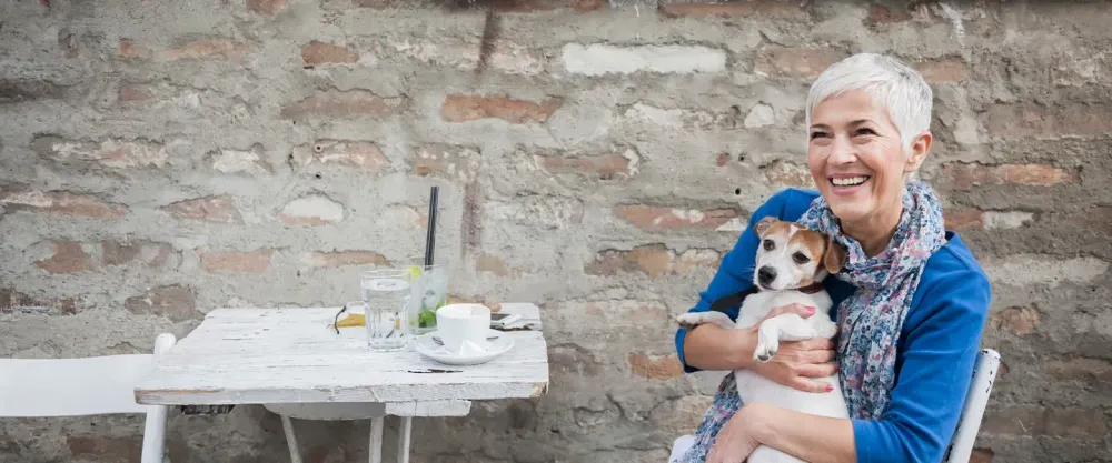 Older white haired woman sitting on a patio and holding a small dog while smiling